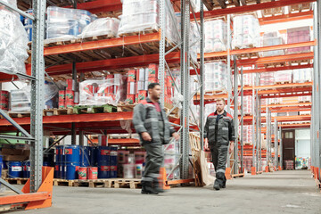 Two young intercultural male workers in uniform moving along huge shelves