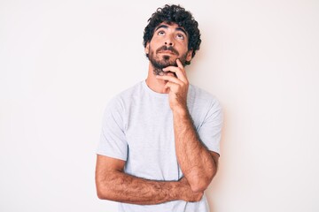 Handsome young man with curly hair and bear wearing casual tshirt serious face thinking about question with hand on chin, thoughtful about confusing idea