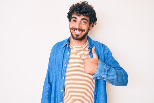 Handsome young man with curly hair and bear wearing casual denim jacket doing happy thumbs up gesture with hand. approving expression looking at the camera showing success.