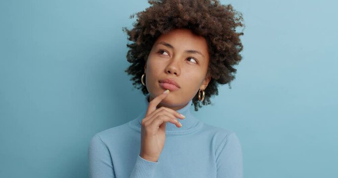 Serious dark skinned woman with thoughtful expression keeps finger on face concentrated somewhere pensively has curly bushy hair dressed in poloneck isolated over blue background. Thoughts concept
