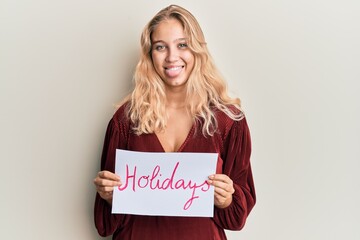 Fototapeta na wymiar Young blonde girl holding holidays banner message sticking tongue out happy with funny expression.