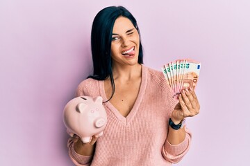 Young caucasian woman holding euro banknotes and piggy bank winking looking at the camera with sexy...