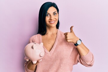 Young caucasian woman holding piggy bank smiling happy and positive, thumb up doing excellent and approval sign