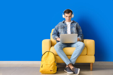 Male student with laptop studying online near color wall