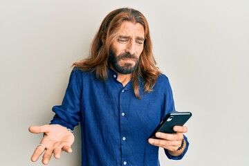 Attractive man with long hair and beard using smartphone clueless and confused expression. doubt...