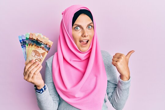 Young caucasian woman wearing islamic hijab holding canadian dollars banknotes pointing thumb up to the side smiling happy with open mouth