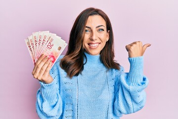 Young brunette woman holding 20 israel shekels banknotes pointing to the back behind with hand and...