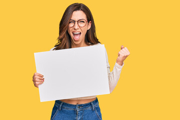 Fototapeta na wymiar Young brunette woman holding blank empty banner screaming proud, celebrating victory and success very excited with raised arms