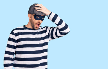 Young handsome man wearing burglar mask surprised with hand on head for mistake, remember error....