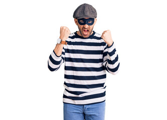 Young handsome man wearing burglar mask angry and mad raising fists frustrated and furious while...