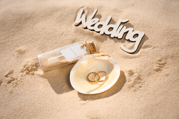 Fototapeta na wymiar Shell with gold rings, invitation in glass bottle and word Wedding on sandy beach