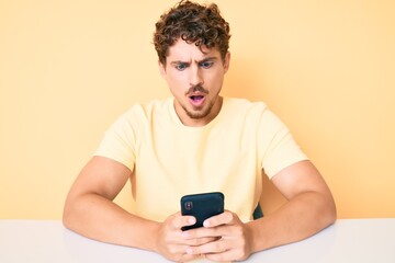 Young caucasian man with curly hair using smartphone sitting on the table scared and amazed with open mouth for surprise, disbelief face