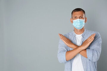 Man in protective mask showing stop gesture on grey background, space for text. Prevent spreading of coronavirus