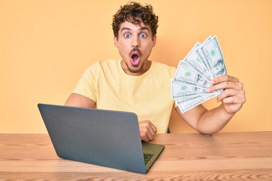 Young caucasian man with curly hair working using computer laptop holding usa dollars banknotes scared and amazed with open mouth for surprise, disbelief face