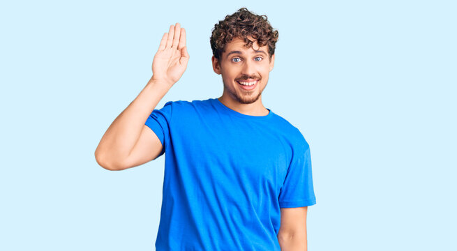 Young handsome man with curly hair wearing casual clothes waiving saying hello happy and smiling, friendly welcome gesture
