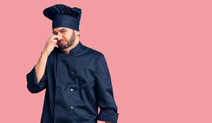 Young hispanic man wearing cooker uniform smelling something stinky and disgusting, intolerable smell, holding breath with fingers on nose. bad smell