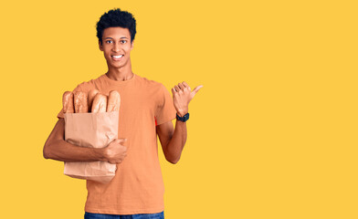 Young african american man holding paper bag with bread pointing to the back behind with hand and thumbs up, smiling confident