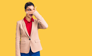 Young beautiful woman wearing business shirt and glasses peeking in shock covering face and eyes with hand, looking through fingers with embarrassed expression.