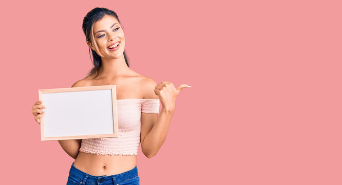 Young beautiful woman holding empty white chalkboard pointing thumb up to the side smiling happy with open mouth