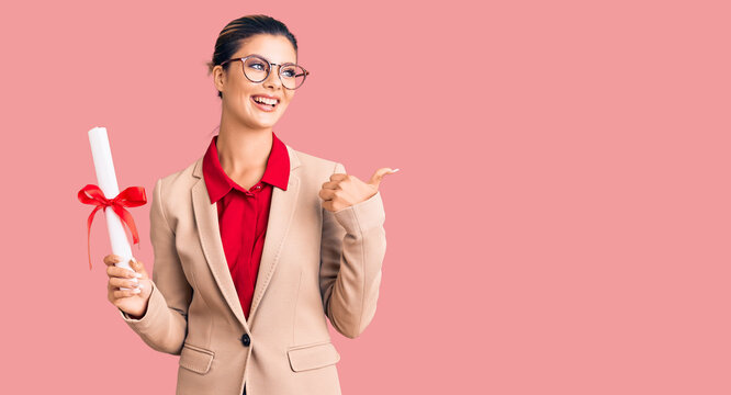Young beautiful woman wearing glasses holding graduate degree diploma pointing thumb up to the side smiling happy with open mouth