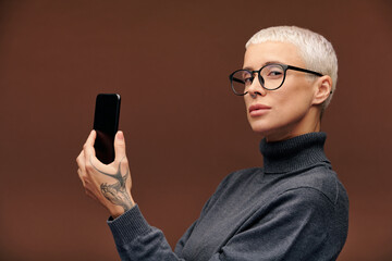 Young serious blond businesswoman in pullover and eyeglasses holding smartphone