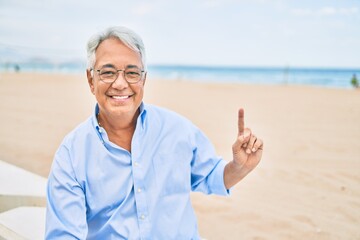 Handosme hispanic man with grey hair smiling happy at the beach, enjoying holidays pointing with finger at copy space