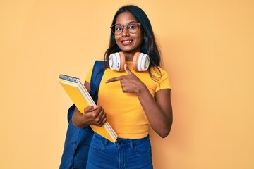 Young indian girl holding student backpack and books smiling happy pointing with hand and finger