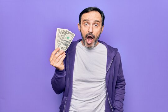 Middle age handsome man holding bunch of dollars banknotes over isolated purple background scared and amazed with open mouth for surprise, disbelief face