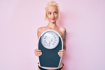 Young blonde woman with tattoo standing shirtless holding weighing machine smiling looking to the side and staring away thinking.