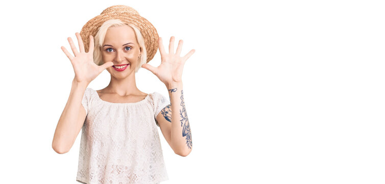 Young blonde woman with tattoo wearing summer hat showing and pointing up with fingers number ten while smiling confident and happy.