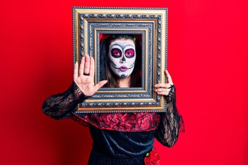 Young woman wearing day of the dead costume holding empty frame with open hand doing stop sign with serious and confident expression, defense gesture