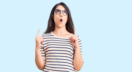 Brunette teenager girl wearing casual clothes and glasses amazed and surprised looking up and pointing with fingers and raised arms.