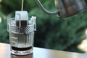 Pouring hot water into glass with drip coffee bag from kettle on wooden table, closeup