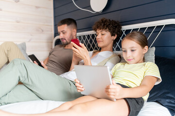 Little girl in casualwear using tablet while sitting on bed next to her parents