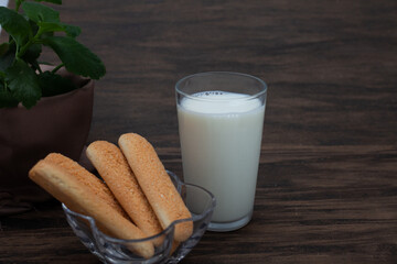 Brazilian champagne biscuit in focus ahead with glass of milk and vase with green leaves slightly defocused on wooden background. Front view with clean space for design.