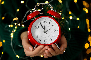 Woman holding alarm clock against blurred lights, closeup. New Year countdown