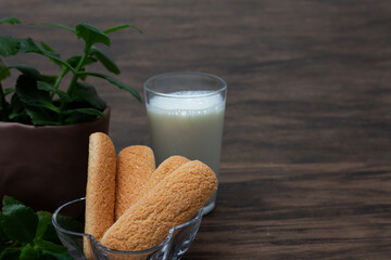 Brazilian champagne biscuit in focus ahead with glass of milk and vase with green leaves slightly defocused on wooden background. Front view with clean space for design.