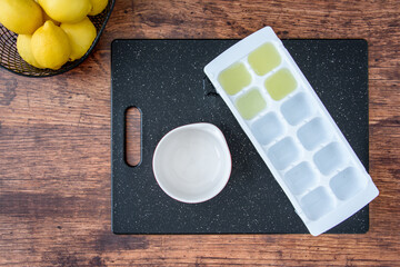 Fresh lemon juice in an ice cube tray, black cutting board, wood table, basket of lemons, white...