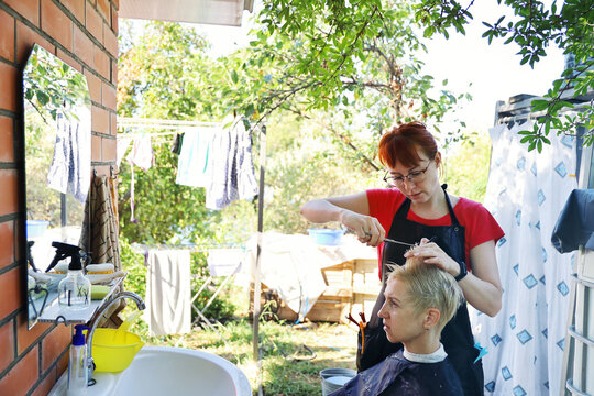 Young woman, professional hairdresser makes haircut to another person outdoors in the yard in the household part, not in the beauty salon. New reality in pandemic
