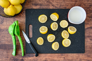 Fresh lemon juice in a white bowl, lemon halves on a black cutting board, paring knife, wood table,...