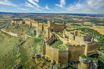 Aerial view of Gormaz castle in Spain with dozens of square projecting towers, loopholes, inner and outer bailey on a bare mountain top near the Duero river with dreamy blue sky