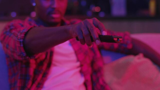 Close-up of casual black man in shirt lying on hte sofa pressing buttons of remote control watching TV in stylish neon interio0r living room apartment concept.
