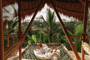 Young pretty woman sit on hammock on balcony of a bamboo eco house in the jungle. Eco tourism on Bali island