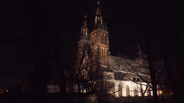 The illuminated gothic medieval church of saint Peter and Paul with its two spires at Vy&scaron;ehrad,Prague,Czechia,at night,seen through silhouettes of leafless tree branches,looking dark and ominous.