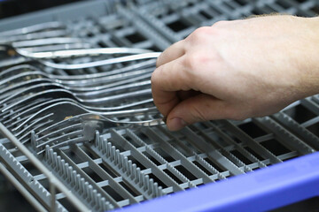 A man loads dirty dishes, plates, spoons, forks, cutlery into dishwasher tray.