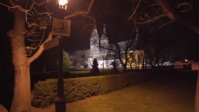 The illuminated gothic medieval church of saint Peter and Paul with its two spires at Vy&scaron;ehrad,Prague,Czechia,at night,behind a park with trees, a hedge,a street lantern with a sign and statues,zoom.