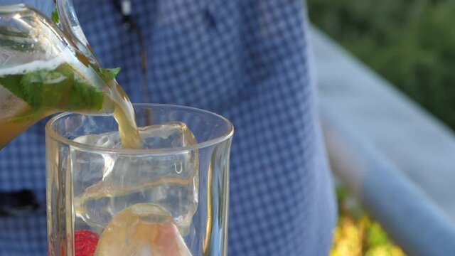 Slow motion pour of fruity orange alcoholic drink with leaf and raspberry from clear jug into ice filled glass filling up on summer day.