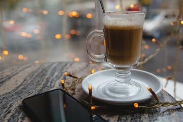 A glass of coffee on a saucer on the table and a smartphone on the background of a window with bokeh
