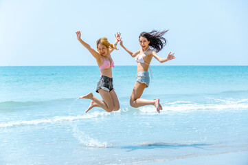 Happy two beautiful Asian woman friend tourist playing and jumping together on the beach in summer day. Happy pretty girl relax and enjoy outdoor lifestyle in summer holiday vacation