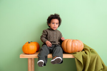 Fototapeta premium Cute African-American baby girl with pumpkins sitting on bench near color wall